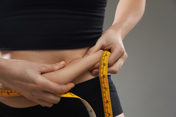 Woman with measuring tape touching belly fat on grey background, closeup. Overweight problem