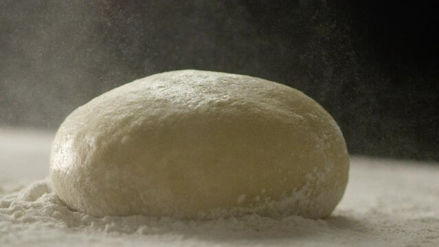 Person throwing flour over ball of dough on counter black background