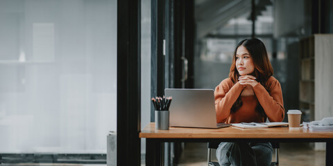 Young businesswoman with hands clasped, sitting at her desk in a modern office, deep in thought. - obrazy, fototapety, plakaty