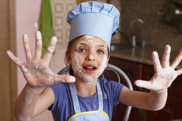 5 year old funny girl in blue dress and dressed as chef in kitchen laughs and showing her fingers...
