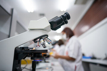 A woman is wearing a white lab coat and is looking through a microscope