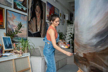 Focused inspired female artist drawing on canvas in art studio with houseplants. Interested young woman painter holding colour palette and paintbrush applies oil paint to complete painting, on easel.