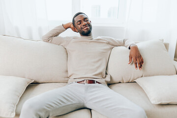 Relaxed African American Man Sitting on a Modern Sofa, Smiling and Holding a Smartphone, Enjoying a...