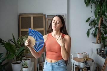 Exhausted woman suffers from heat fanning with paper fan. Stressed overheated girl sweating...