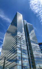 Obraz premium Dominating the frame, a modern skyscraper ascends towards the clear blue sky, lined with fluffy white clouds. Its glass facade reflects the surrounding environment, creating a play of light and shadow