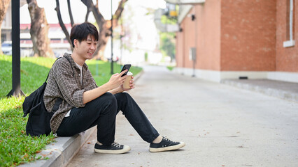 A happy Asian male college student sits on the street in a campus, using his smartphone.