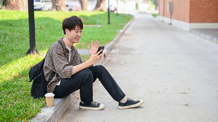A cheerful Asian student sits on the street talking with his friend on a video call on his phone.