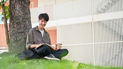 A happy, relaxed Asian male college student enjoys reading a book under the tree in the campus park.