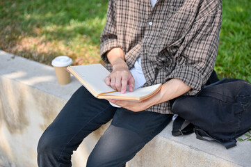 A cropped image of a male college student sits on a stone bench in a park, reading a book outdoors.
