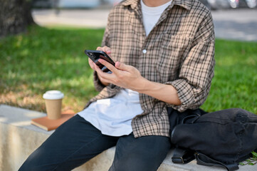 A cropped image of a male college student sits on a stone bench in a park, using his smartphone.