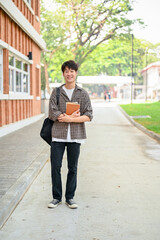A positive Asian man college student, stands in front of a brick building on campus, holding books.