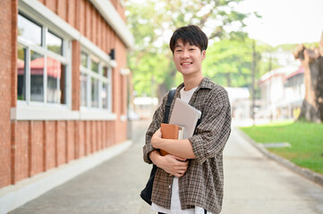 A cheerful Asian man student stands outside a brick building on campus, holding a laptop and books.