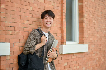 A smiling young Asian male college student stands by a brick wall on campus, carrying a backpack.
