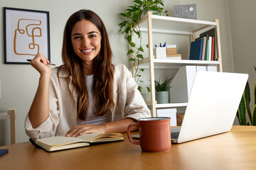 Young happy female caucasian entrepreneur handwriting on a notebook working at home office looking...