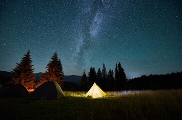 Night camping in mountains under starry sky. Tourist tents in campsite near burning campfire under...