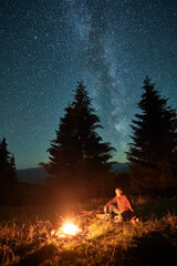Night camping in mountains under starry sky with Milky way. Man sitting near campfire, holding...