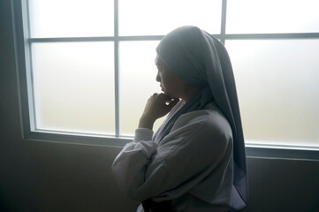 Back view of young muslim woman standing near window with sad expression 