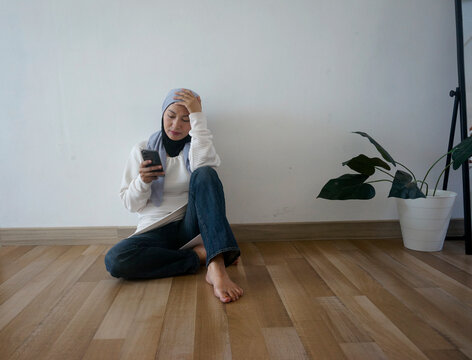 Full length image of Asian woman sitting on floor and touching head while looking at mobile phone, feeling sad and confused