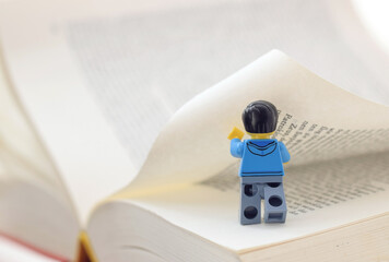 Naklejka premium Lego minifigure of cute man is turning a page in book. Concept image to popularity of reading. Editorial illustrative image of education and learning.