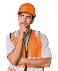 Young Hispanic worker in safety gear looking sideways with doubtful and skeptical expression.
