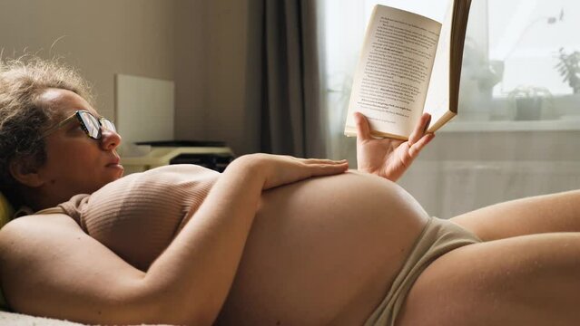 A pregnant woman lying cozily on a bed and reading a book. The woman is wearing a beige top. With her left hand she holds a book over her belly, while her right hand gently strokes her belly