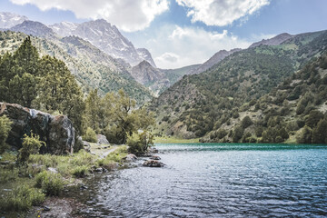 Mountain panorama, landscape with rocky peaks and blue turquoise lake Chukurak in the Fan Mountains in Tajikistan, hills covered with forest on a sunny summer day