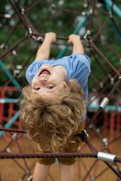 The child is hanging upside down on the playground equipment. The outdoor playground for children in summer park. Kid play on playground under the tree. Excited kid doing rock climbing.