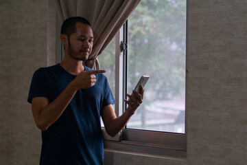 feeling happy of Young Asian man in blue t-shirt  and pointing to smart phone beside of window