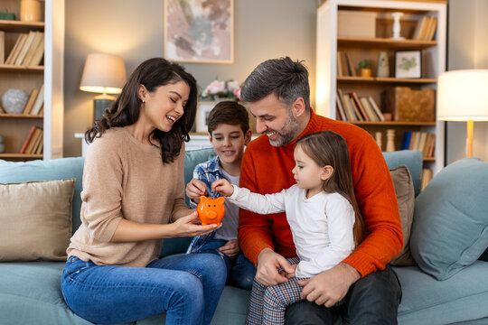 Happy family cheerful mother and father with kids smiling and putting coins into piggy bank while sitting on sofa at home