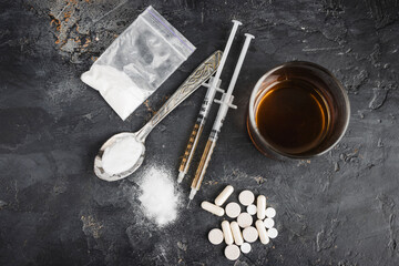 Narcotic substances in spoon, dope powder in transparent plastic bag, syringes with drugs dose, white pills and glass of alcohol drink on dark textured background, top view