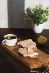 Sandwich with sliced ham butter and cheese on wooden cutting board near mug of tea with lemon