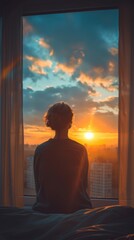 A girl is sitting on a bed in front of the window and looking at the sunset.