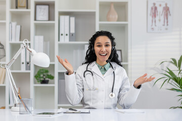 A cheerful female doctor in a white coat is engaged in a virtual consultation, communicating warmly with a patient, sitting in a modern office.