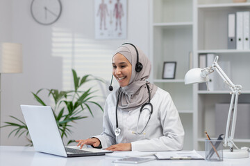 A professional and cheerful female doctor in a hijab provides online consultations from her well-organized office, using a laptop and headset.