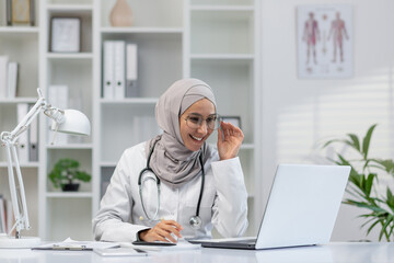 A female healthcare professional in hijab smiling while working on her laptop, probably engaged in an online consultation. Well-lit modern office environment.