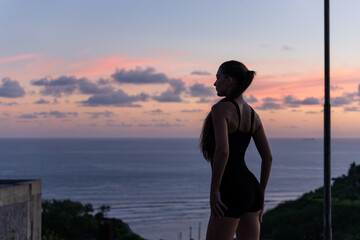 Fototapeta premium Sunset Gaze Profile view of a young woman in a black bodysuit looking out to the ocean at sunset, with dramatic clouds in the sky