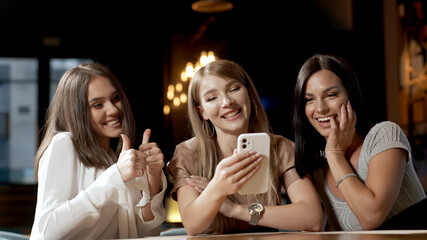 Girlfriends take a selfie on a mobile phone camera while sitting in a cafe. Three attractive girls are photographed with a smartphone camera.