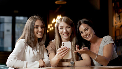A smiling woman and her beautiful friends in a cafe where they are meeting. A woman shows something on her smartphone. Women discuss each other.