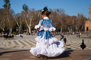 a little girl dancing flamenco dressed in a white dress with ruffles and blue fringes in a famous...