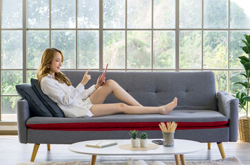 Young asian woman in white shirt and short pant, sitting on a sofa, using a tablet and smiling, relax in a bright room with large window.