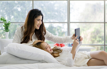 Young asian couple in bedroom with a bright, natural light through the window. Smiling while holding tablet computer, taking a selfie or video call. Moment of relaxation and connection