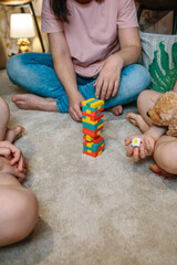 Unrecognizable family having fun making a tower with wooden stacking piece game at home. Mother and sons enjoying together in leisure time. Happy little girl holding a dice and stuffed teddy dog.