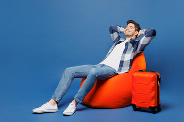 Traveler smiling happy man wear casual clothes sit in bag chair near suitcase bag isolated on plain blue background. Tourist travel abroad in free time rest getaway. Air flight trip journey concept.