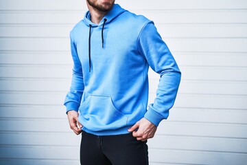 A bearded man in a blue hoodie leans against a white wall. This is a mockup for logo and brand swag...