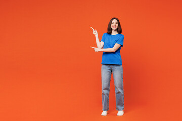 Full body young smiling cheerful fun cool happy woman she wear blue t-shirt casual clothes point index finger aside on area isolated on plain red orange background studio portrait. Lifestyle concept.