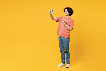 Full body young happy man wear pink t-shirt casual clothes doing selfie shot on mobile cell phone post photo on social network show v-sign isolated on plain yellow orange background Lifestyle concept