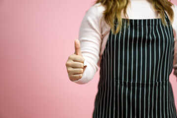 Young woman in pink sweater and striped apron give a thumbs up gesture. Portrait on pink background with studio light.