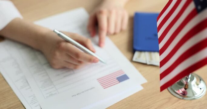 Woman fills in Travel visa application form at desk with American flag. Lady signs official documents sitting at wooden table slow motion