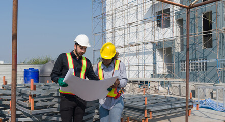 Two construction worker looking at a floor plan, standing in front of a large building site with helmet and safety gear. Day time work safety check. Work environment at the site of housing projects.