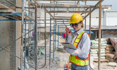 A builder in safety uniform standing at a building site, holding a walkie-talkie communicating with other team member. Tablet in her hand used for review construction plan or checking schedule.